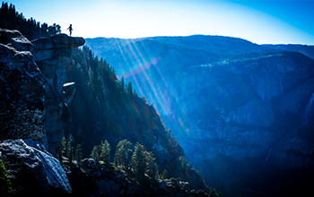 A woman stands on the top of a cliff, looking over the mountains